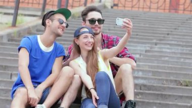 Canlı bir üçlü genç arkadaş, iki erkek ve bir kadın, açık hava basamaklarında oturup selfie çekerken ışıldıyorlar. Kaygısız kahkahaları ve canlı kıyafetleri yaz eğlencesinin ve arkadaşlığın özünü yansıtıyor. Sıcak anılar yaratıyor.