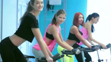 Canlı bir spor salonu ortamında zarif bir kız sabit bir bisiklete güvenle pedal çeviriyor ve kameraya gülümsüyor. Arkasında renkli spor kıyafeti giymiş bir grup atletik kadın enerjik bir şekilde gazla dolu canlı bir atmosfer yaratıyor.