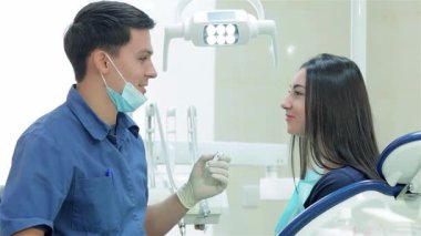 Mavi üniforma ve eldiven giyen dost canlısı bir diş hekimi, genç bayan hastasına yapılacak tedaviyi dikkatle açıklarken diş aletini elinde tutuyor. Parlak, klinik ortam, kız olarak bakım ve profesyonellik atmosferini arttırır.