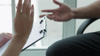 Endişeli bir doktor, hasta ellerini kaldırıp konuşmalarının duygusal ağırlığını vurgulayan bir jest yaparken dikkatlice dinler. Parlak klinik ayarı, pencereden içeri doğru akan doğal ışıkla,