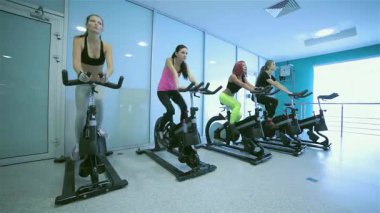 Modern bir spor salonunda, bir grup arkadaşın durağan bisikletler üzerinde enerjik bir şekilde pedal çevirdiği ve dikkatlerinin ileri yöneldiği hareketli bir sahne sergileniyor. Renkli spor giysileri içinde, bu atletik bireyler kararlılık ve dostluk yayıyorlar, somutlaşıyorlar.