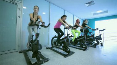 Hareketli bir grup sporcu enerjik bir bisiklet seansına katılıyor, parlak, iyi ışıklandırılmış bir spor salonunda hareketsiz bisikletlerle pedal çeviriyorlar. Renkli spor kıyafetleri içinde bu fitness meraklıları kararlılıklarını ve dostluklarını sergiliyorlar.
