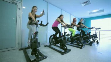 Parlak bir spor salonunda dinamik bir sahne sergileniyor. Sportif kızlar hareketsiz bisikletlerle pedal çeviriyor, başparmakları parlıyor ve gülümsüyor. Hareketli spor kıyafetleri ve yoldaşlıkları ilham verici bir atmosfer yaratıyor, fitness ve dostluk gösterisi yapıyorlar.