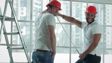 Parlak, havadar bir dairede, turuncu kasklı iki yetenekli işçi pencereleri bant, takım çalışması ve uzmanlığıyla ölçüyorlar. Gülümsemeleri güven saçıyor. Tadilata hazırlanıyorlar. İnşaat ve işbirliği ruhunu somutlaştırıyorlar.