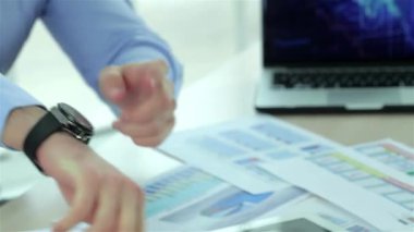 Dinamik bir iş ofisinde, yakın plan bir iş adamını saatini işaret ederek aciliyet ve odaklanma sinyali vererek yakalar. Resmi giyinmiş iki başarılı iş adamı dizüstü bilgisayar ve çizelgelerle, profesyonelliği ve yüksek bahislerde takım çalışmasını somutlaştırıyor.