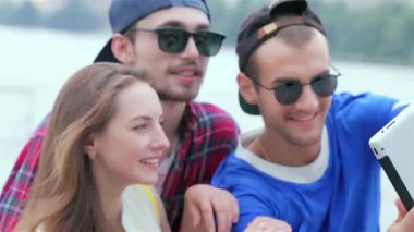 Üç neşeli arkadaş, bir erkek ve iki kadın, suyun kenarında selfie çekmek için birlikte poz veriyorlar, mutluluk ve dostluk saçıyorlar. Şık güneş gözlükleri ve canlı kıyafetleri yazın kaygısız ruhunu yansıtıyor. Bu anı mükemmel bir enstantane haline getiriyor.