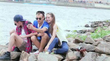 Üç genç arkadaş nehir kenarındaki güneşli kayalara oturup gülüyor ve selfie çekiyor ve güzel anılar yaratıyorlar. Kaygısız gülümsemeleri ve enerjik enerjileri güzel bir yaz gününde arkadaşlığın özünü yayıyor.