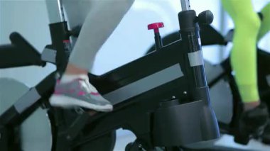 Yakın çekim, hareketli bir spor salonu atmosferiyle çevrili hareketsiz bisikletlerle güçlü pedal çeviren atletik ayakların hareketini yakalar. Sahne fitness tutkunlarının kararlılığını ve enerjisini gözler önüne seriyor.
