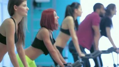 Canlı beş arkadaştan oluşan bir grup enerjik bir bisiklet seansına katılıyor, parlak ve modern bir spor salonunda hareketsiz bisikletlerle pedal çeviriyorlar. Şık spor kıyafetleri içinde odaklanmış ifadeleri ve senkronize hareketleri ilham verici bir şey yaratıyor.