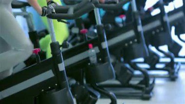 Spor salonundaki canlı bir sahnede bir grup atletik arkadaş hareketsiz bisikletler üzerinde pedal çeviriyor, odaklanmış ifadeleri kararlılığı yansıtıyor. Spor kıyafetlerinin parlak renkleri, özellikle de neon tozluklarının.