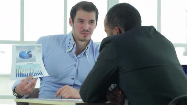 Parlak bir ofis ortamında, iki kendine güvenen işadamı ayrıntılı bir iş planını analiz ederek dinamik bir tartışmaya girişirler. Açık mavi gömlekli bir adam, tutkuyla çizelgeleri sunarken, diğeri, parlak siyah bir takım elbise içinde, dikkatle dinliyor.