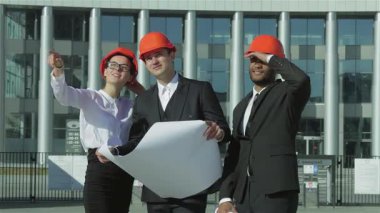 Canlı turuncu miğferler takmış üç kendine güvenen iş mimarı iddialı projelerini incelemek üzere sahada duruyor. Modern mimarinin bir zeminine karşı yenilikçi yapı fikirlerini tartışırken ifadeleri kararlılık ve takım çalışması yayıyor.