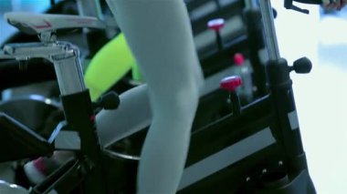 Atletik bir kız, dört formda arkadaşı tarafından çevrili sabit bir bisikletin üzerinde yoğun bir şekilde pedal çeviriyor ve hepsi de yüksek enerjili bisiklet seansına katılıyor. Parlak spor salonu atmosferi motivasyon saçıyor, güçlü bacaklar ve kararlı ifadeler sergiliyor.