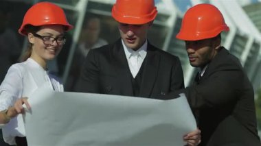 Üç kendine güvenen iş mimarı, bir kadın ve turuncu kasklı iki adam, açık havada durup bir planı dikkatle gözden geçiriyorlar. Odaklanmış ifadeleri ve takım çalışması, yenilikçi yapı fikirlerini hayata geçirmenin heyecanını vurguluyor.