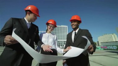 Parlak turuncu kasklı üç kendine güvenen iş mimarı dışarıda duruyor ve hevesle projeleri inceliyor. Takım çalışmaları ve ortak vizyonları, yaklaşan inşaat projesi için yenilikçi fikirleri tartışırken başarıya ışık tutuyor.