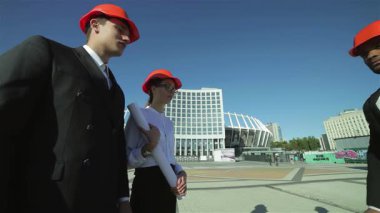 Parlak turuncu miğferler takmış üç kendine güvenen mimar, hareketli bir inşaat sahasında birlikte duruyor ve iddialı projelerini inceliyorlar. Onların odaklanmış ifadeleri ve dinamik takım çalışması yeniliğin özünü vurgular ve