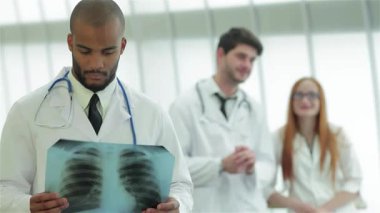 Kendini adamış genç bir doktor, beyaz önlüklü iki destekçi meslektaşı tarafından çevrelenmiş sağlıklı akciğerlerin röntgenini inceliyor. Parlak, havadar klinik kurulumu takım çalışmasının ve profesyonelliğin atmosferini geliştiriyor ve hastalara olan bağlılığını gösteriyor.