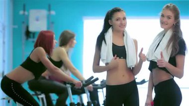 İki kendine güvenen genç kadın canlı bir spor salonunda poz veriyor, kameraya başparmaklarını kaldırırken, arkadaşları arka planda hareketsiz bisikletler üzerinde enerjik bir şekilde pedal çeviriyor. Bu canlı sahne pozitiflik ve dostluk saçıyor, dinamik bir zindelik sergiliyor.