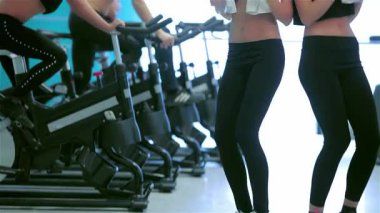 Canlı bir spor salonu ortamında, iki kız aynı neşeli anı paylaşıyor, arkadaşları sabit bisiklete binerken gülüyorlar ve başparmaklarını kaldırıyorlar. Bu canlı sahne, arkadaşlığın özünü ve fitness motivasyonunu yakalar.