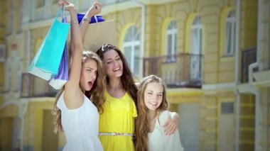 Üç neşeli kız, kollarını kaldırmış canlı alışveriş torbalarıyla, güneşli bir caddede yürürken mutluluk saçıyorlar. Kahkahaları ve kaygısız ruhları bulaşıcı bir dostluk ve eğlence ortamı yaratıyor, özü yakalamak için mükemmel.