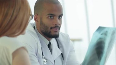 Beyaz önlüklü odaklanmış bir erkek doktor, akciğer sağlığının önemini vurgulayan bir kadın hastaya göğüs röntgenini özenle açıklar. Parlak, modern klinik kurma, profesyonellik ve bakım atmosferini geliştirir.