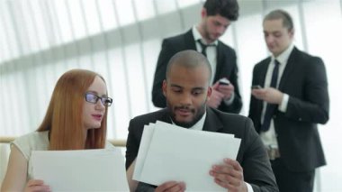 Kızıl saçlı ve gözlüklü kendine güvenen bir kadın, her ikisi de önemli belgeleri inceleyen iyi giyimli bir adamla canlı bir tartışmaya girer. Arka planda, diğer iki profesyonel, bir erkek ve bir kadın, kendi cihazlarına odaklanmış, canlı bir şekilde