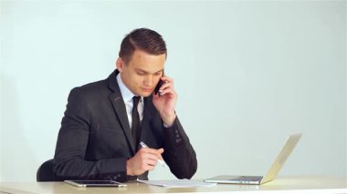 Sivri takım elbiseli genç bir işadamı cep telefonuyla konuşurken kalemle notlar yazıyor, profesyonellik ve kararlılık yayıyor. Bir dizüstü bilgisayar ve tabletle tamamlanmış gösterişli ofis ortamı,
