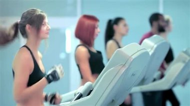 Gösterişli spor kıyafetleri içindeki kararlı genç bir kadın canlı bir spor salonundaki diğer sporcularla çevrili bir koşu bandında koşuyor. Her bir birey sağlık ve zindeliğe olan bağlılığını gösterirken atmosfer enerji ile uğulduyor.
