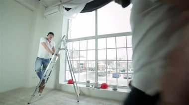 Parlak, havadar bir dairede, yetenekli bir müteahhit, arka planda hızla ilerlerken perdeleri asmaya hazırlanıyor. Sahne, canlı boya kutuları ve güvenlik ile takım çalışmasının ve dönüşümün enerjisini yakalıyor.