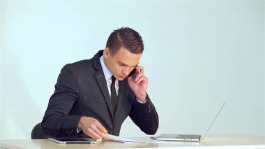 Keskin bir takım elbise giymiş odaklanmış genç bir işadamı, önemli bir telefon görüşmesi yaparken gülümsüyor, masasındaki veri tablolarına bakıyor. Pürüzsüz bir dizüstü bilgisayar ve tabletle tamamlanan modern ofis ortamı profesyonel atmosferi geliştiriyor.