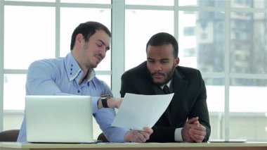 Biri açık mavi gömlekli, diğeri keskin siyah takım elbiseli iki kendine güvenen işadamı, şık bir ofis masasında belgeleri dikkatle inceliyor. Geniş pencereli, aydınlık, modern ofis ortamı, doğal ışığı arttırır.