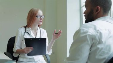 Odaklanmış bir kadın doktor erkek hastasıyla düşünceli bir konuşma yapar, iyi aydınlatılmış bir klinikte sıcak ve profesyonel bir atmosfer sunar. Doktor, steteskopunu boynuna dolamış, notlar alırken dikkatle dinliyor.