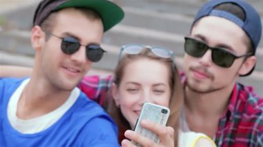 Neşeli bir kız iki arkadaşıyla eğlenceli bir selfie çeker. Hepsi şık bir güneş gözlüğü takar ve parlak bir şekilde gülümserler. Bu hayat dolu an, güneşli bir günde gülüşmeler ve dostlukla dolu kaygısız gençlik ruhunu gösteriyor.