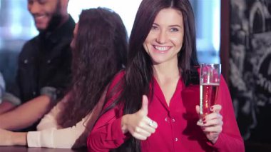 Canlı bir arkadaş grubu, coşkulu bir barda, elinde bira tutarken sevinçle baş parmak kaldıran genç bir kadınla kutlama yapıyor. Kahkahalar havayı doldururken atmosfer heyecan verici ve herkes favori futbol takımı için tezahürat yapıyor.