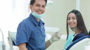 Mavi üniforma ve eldiven giyen neşeli bir dişçi, gülümseyen bir kadın hasta dişçi koltuğunda rahatça otururken aletleri hazırlar. Kliniğin parlak, cezbedici atmosferi bakım ve profesyonellik duygusunu geliştirir, diş hekimliği yapar.