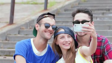 Genç arkadaşlardan oluşan canlı bir üçlü, mutluluk saçıyor, açık havada birlikte poz veriyorlar, akıllı telefonlarıyla selfie çekiyorlar. Neşeli ifadeleri ve şık kıyafetleri kahkaha ve arkadaşlıkla dolu tasasız bir yaz gününü yansıtıyor.