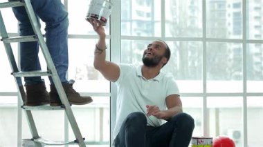 Yerde oturan neşeli bir adam merdiven üzerinde çalışan bir işçinin tuttuğu boya kutusuna hevesle uzanır, takım çalışmasını ve ev gelişimindeki işbirliğini somutlaştırır. Bu canlı sahne, yenilenmenin özünü yakalıyor.