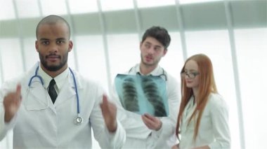 Başarılı bir erkek doktor, ekibi sağlıklı akciğerlerin röntgenini incelerken gururla iki parmağını kaldırır. Bu dinamik sahne, parlak ve çağdaş bir klinikte takım çalışmasının ve uzmanlığın özünü yakalar, sağlığın ve
