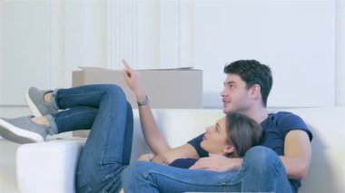 Taşınan kutularla dolu sakin, boş bir dairede, sevgi dolu bir çift yeni başlangıçları için mutluluk ve heyecan saçan rahat bir kanepede sıcak bir kucaklaşmayı paylaşıyor. Rahat duruşları ve gülümsemeleri yeni bir başlangıç yapmanın hazzını yansıtıyor.