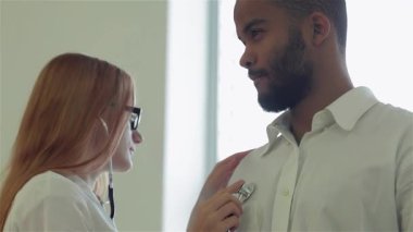 Parlak ve modern bir klinikte odaklanmış bir doktor, genç bir adamın kalp atışlarını dinlemek için steteskop kullanır ve kapsamlı bir muayene sağlar. Bakım etkileşimi, sağlık hizmetlerinde iletişimin önemini vurguluyor.