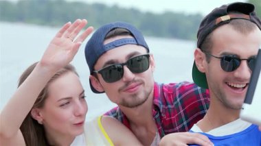 Üç neşeli arkadaş, şık güneş gözlüğü takıyorlar, suyun kenarında eğlenceli bir selfie için poz veriyorlar, mutluluk ve dostluk saçıyorlar. Bu güzel yaz gününde unutulmaz anılar yaratırken kahkahaları havayı dolduruyor.