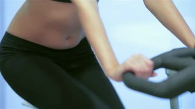 Hareketli bir spor salonu ortamında, atletik bir kız hareketsiz bir bisikletle pedal çeviriyor, fitness 'e olan bağlılığını gösteriyor. Yakın plan karnı ve güçlü bacakları ön plana çıkarıyor. Dört kız arkadaşı birbirlerine girerken takım çalışmasının ruhunu yansıtıyor.