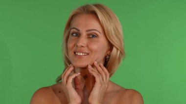 Çekici, olgun bir kadın dalgalı sarı saçları ve büyüleyici mavi gözleri ile kameraya ışıl ışıl gülümsüyor, parlak yeşil renkli bir arka plana hafifçe yüzünü okşuyor. Bu görüntü güzelliğin ve özgüvenin özünü temsil ediyor.