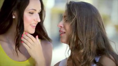 Biri uzun siyah saçlı, diğeri dalgalı kahverengi bukleli iki neşeli kız canlı bir alışveriş bölgesinde gezinirken heyecanlı bir şekilde fısıldaşıyorlar. Neşeli ifadeleri ve renkli kıyafetleri mutluluğu yansıtıyor.