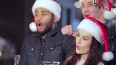 Noel Baba 'nın şen şapkalarıyla süslenmiş, hareketli bir barda futbol takımı için coşkulu bir tezahürat yapan üç arkadaş. Neşeli ifadeleri ve kadeh tokuşturmaları arkadaşlığın ve tatil ruhunun özünü yansıtıyor.