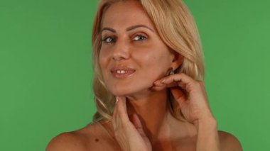 Çekici, olgun, dalgalı sarı saçlı bir kadın canlı, renkli bir arka plana karşı zarifçe poz verir. Yumuşak gülüşü kendine güven ve cazibe saçar. Kusursuz cildini nazikçe okşar, güzelliğin, kadınlığın ve özgüvenin özünü somutlaştırır.