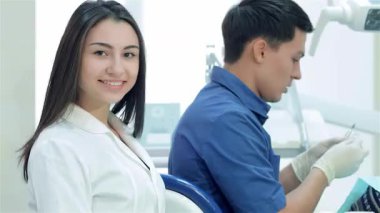 Gıcır gıcır beyaz üniformalı neşeli bir kız-asistan modern bir diş kliniğinde olumlu bir ışık saçar. Arkasında, baş diş hekimi yoğun bir şekilde bir hastaya odaklanıyor. Diş bakımında takım çalışması ve profesyonellik sergiliyor.