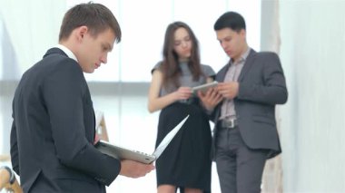 Modern bir ofis ortamında, odaklanmış genç bir adam notlarını incelerken arka planda genç bir çift bir tablet üzerinde görüşlerini paylaşıyor. Bu canlı sahne takım çalışmasının ve yeniliğin özünü yakalıyor, enerjiyi ve