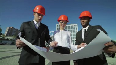 Üç kendine güvenen şık takım elbiseli ve parlak turuncu kasklı mimar dışarıda dikilip ayrıntılı bir iş planını inceliyorlar. Odaklanmış ifadeleri ve işbirlikçi ruhları yenilikçi fikirleri gerçeğe dönüştürmenin heyecanını vurguluyor.