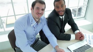 Dinamik bir iş ikilisi, biri açık mavi gömlekli, diğeri keskin siyah takım elbiseli, parmaklarını parlak bir masada, çizelgeler ve dizüstü bilgisayarla süslerken neşe dolu ışıldıyor. Onların kendine güvenen gülümsemeleri pozitiflik, somut takım çalışması ve